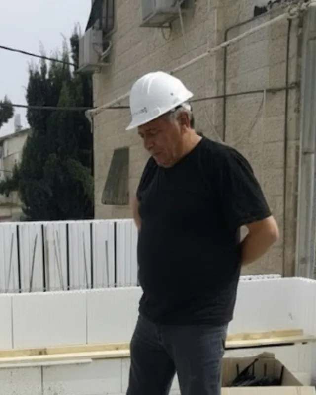 אהרון אלה ז״ל עם קסדה לבנה באתר בנייה, על רקע בניין אבן